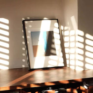 Black aluminum framed artwork on a wooden surface with sunlight casting shadows