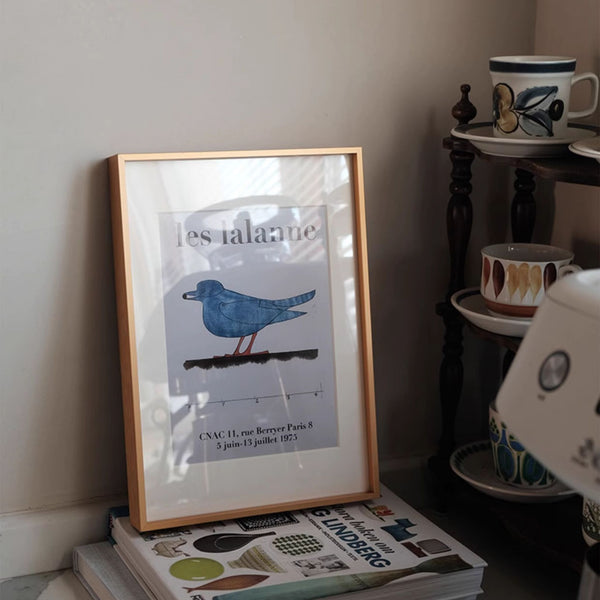 Framed poster with a blue bird illustration on a shelf with books and decorative items.