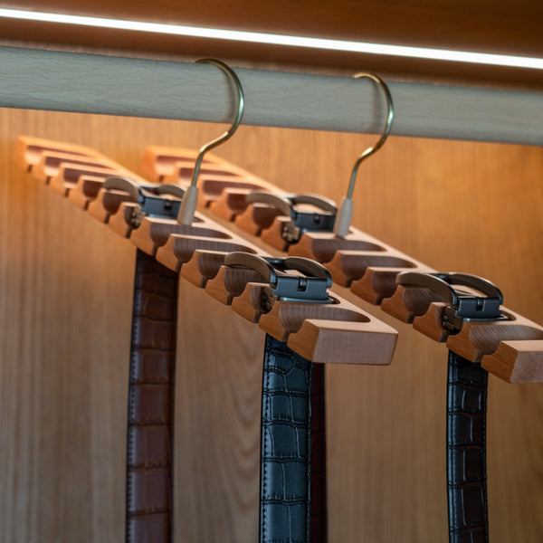 Row of belt hangers with belts on a wooden rack against a wooden wall.