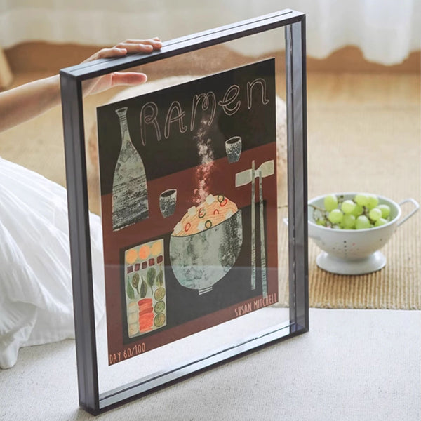 Black acrylic framed artwork titled 'Raven' by Susan Mitchell on a table with a bowl of grapes in the background.