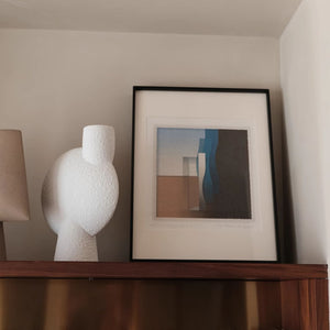 Black Aluminum alloy picture frame with abstract art on a shelf with a textured white sculpture and lamp.