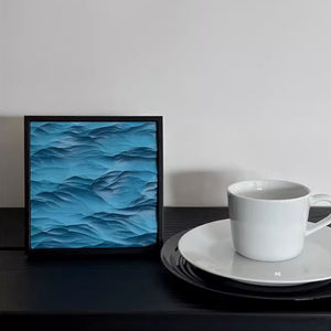 Thin black framed artwork with a 3D wavy blue design next to a white mug on a plate