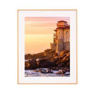 Metal framed artwork of a lighthouse on rocks with a sunset sky.
