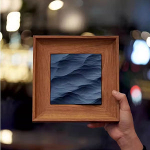 Hand holding a wooden-framed artwork with a blue abstract design against a blurred background