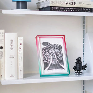 Red and green acrylic framed artwork on a shelf with books and decorative items