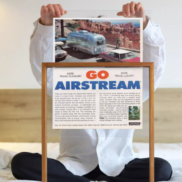 A person holding a teak photo-framed Airstream travel trailer poster with a scenic background