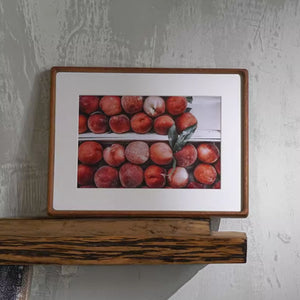 Teak framed picture of peaches on a wooden shelf against a textured wall.