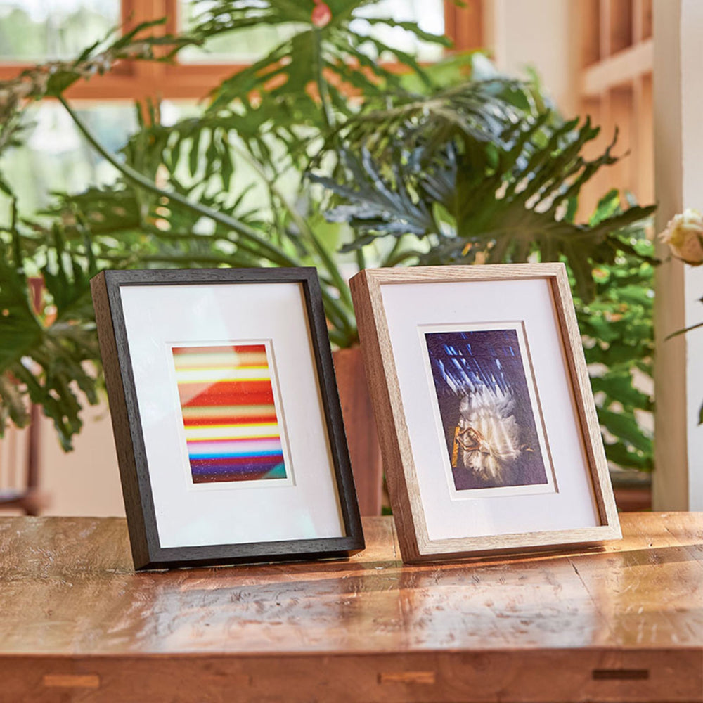 Two framed artworks on a wooden surface with plants in the background. Wenge and white ash wood frame.