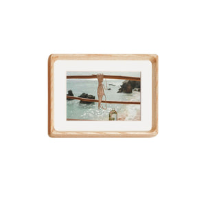 White ash framed artwork of a woman in a swimsuit by the water on a white background