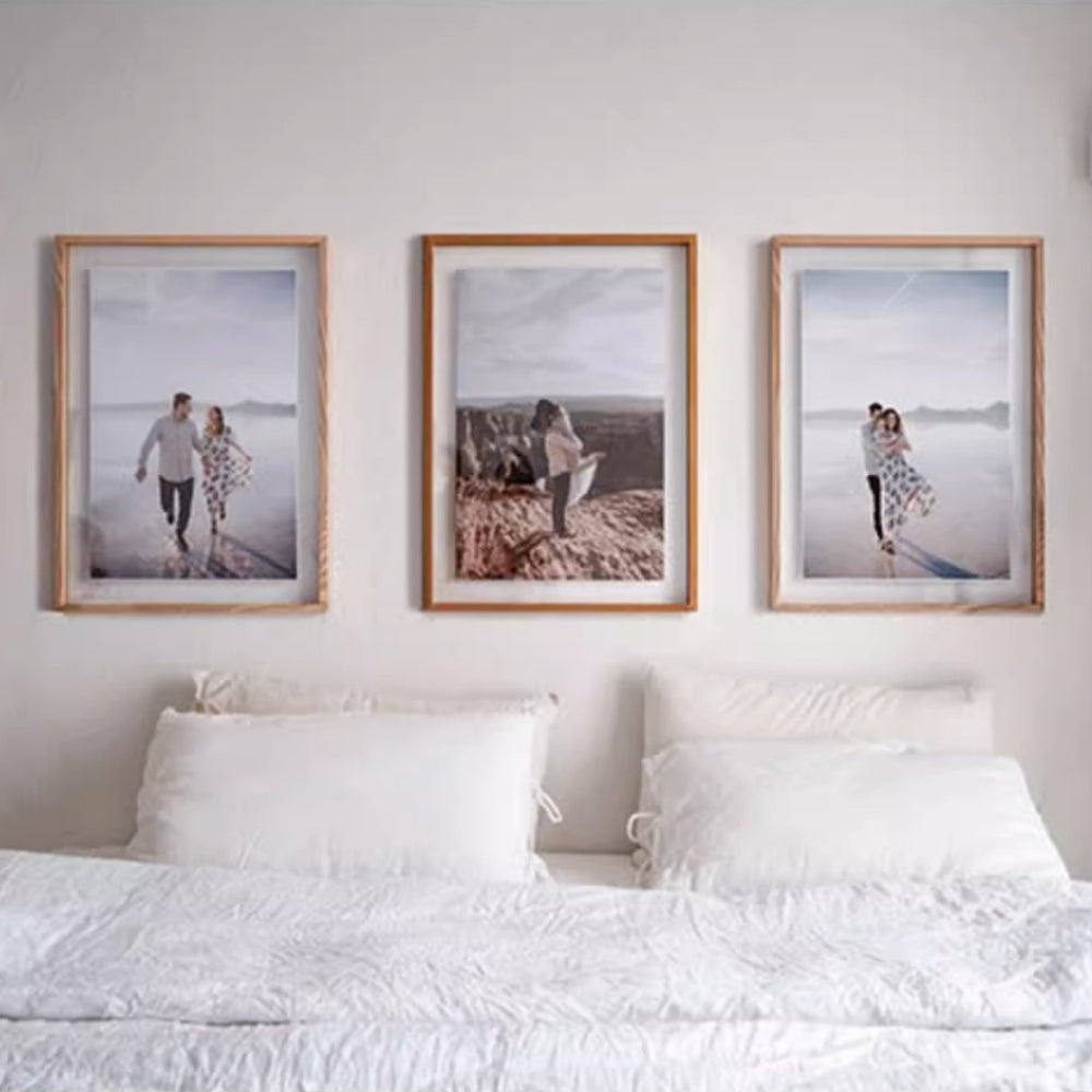 Three framed photographs of a couple on a beach above a bed with white bedding. Two white ash wood plus one teak wood.