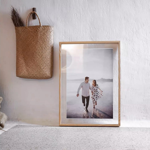 Framed photograph of a couple on a beach with a woven basket above it on a white wall, in a white ash wood picture frame.