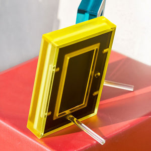 Yellow acrylic picture frame showing the back of the frame and how the plastic peg stands on a red surface with a blurred background