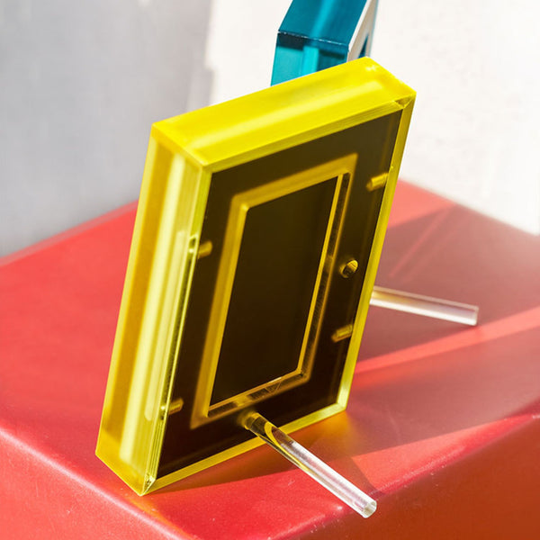 Yellow acrylic picture frame showing the back of the frame and how the plastic peg stands on a red surface with a blurred background