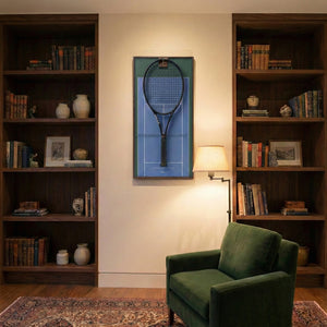 Elegant home library interior showcasing the ONE MAAY US Open hard court tennis racket display mounted on the wall next to a built-in wooden bookshelf and a green reading chair.